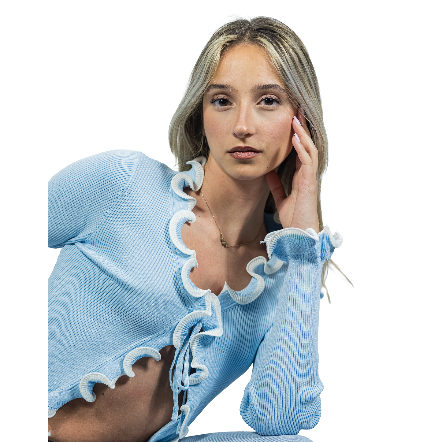 A young woman with long blonde hair wears the MAGLIA WAVE, a light blue crop top with a two-tone wavy hem, resting her face on her hand and looking at the camera against a white background.